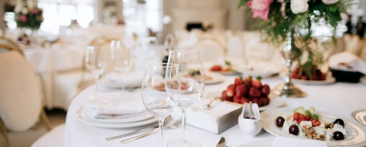 An elegant wedding banquet room with white linens, chairs and flowers with fresh fruits, wine glasses, white plates and wine glasses on a round table.